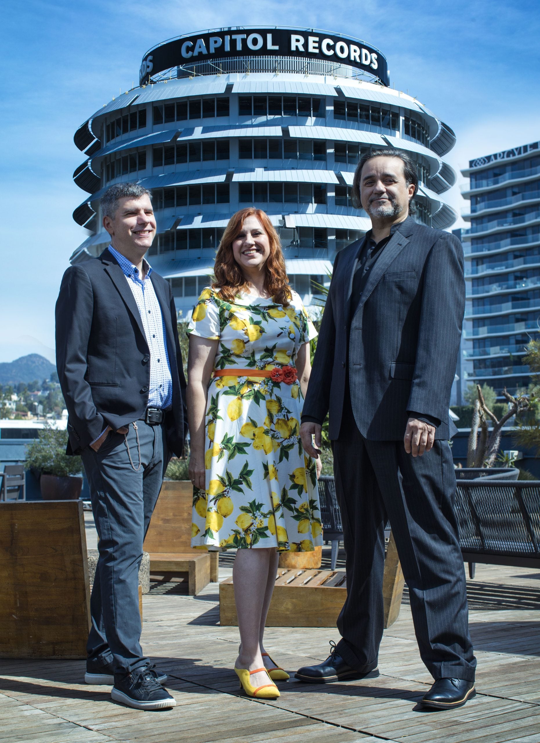 Three people stand outdoors in front of the Capitol Records building, dressed in business and semi-formal attire, posing for a photo under a clear blue sky.