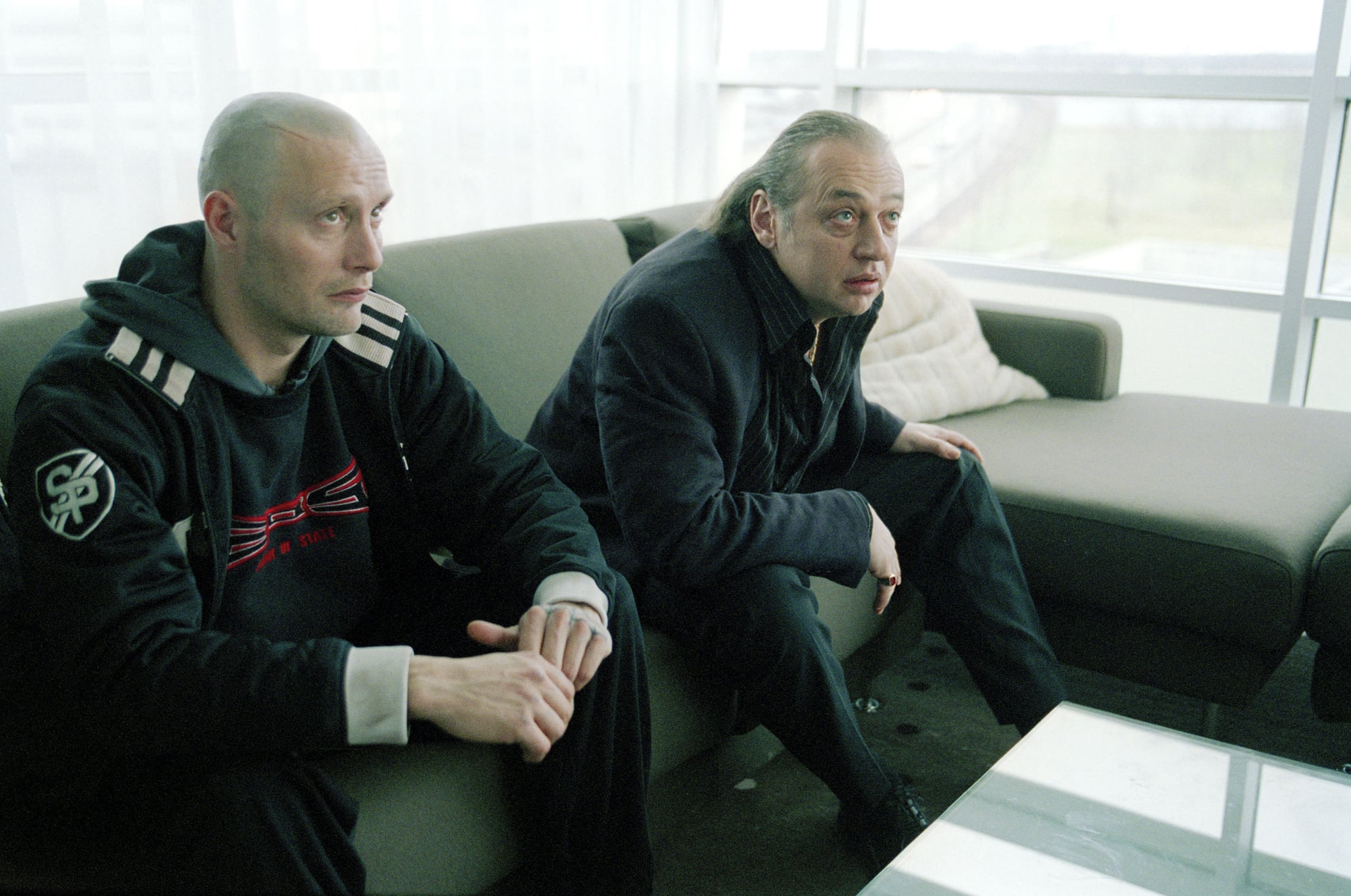 Two men sit on a couch in a modern, light-filled room, looking forward with serious expressions.