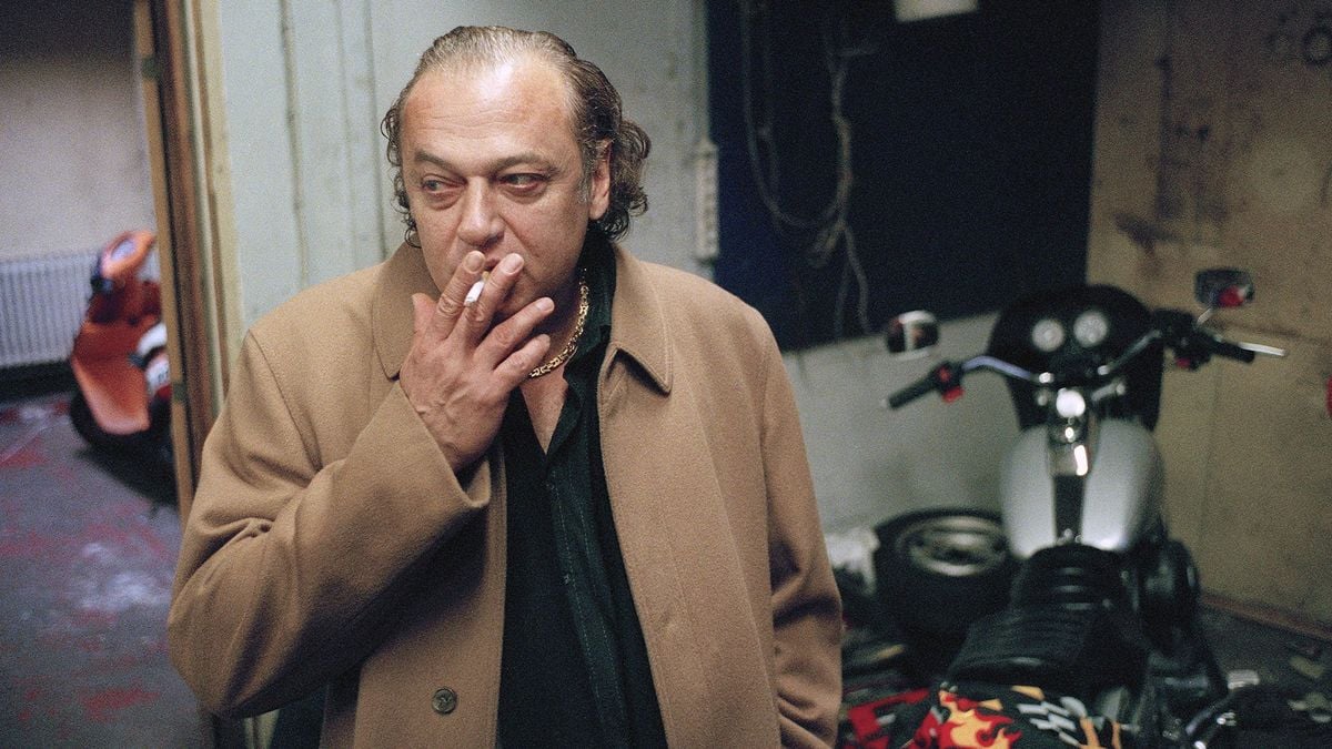 A man with a receding hairline and tan coat smokes a cigarette indoors, standing next to a motorcycle in a dimly lit room.