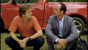 Two men sit squatting in front of a red pickup truck, engaged in a serious conversation outdoors. One wears a red polo shirt, the other a gray suit.