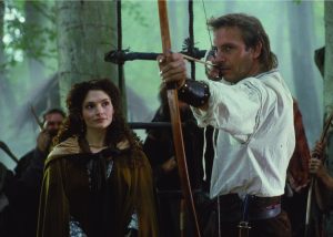 A man aims a bow and arrow while a woman in a cloak stands beside him in a forest setting with several people in the background.