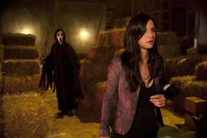A woman in a leather jacket stands in a dimly lit barn with hay bales, while a masked figure in a black robe approaches from behind.