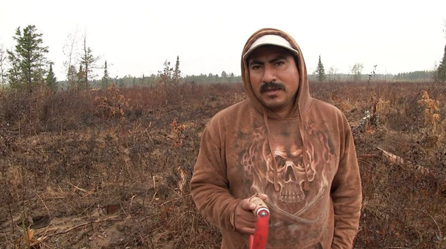 A man in a brown hoodie with a skull design holds a red-handled tool, standing in a barren, cleared outdoor area with sparse trees in the background.