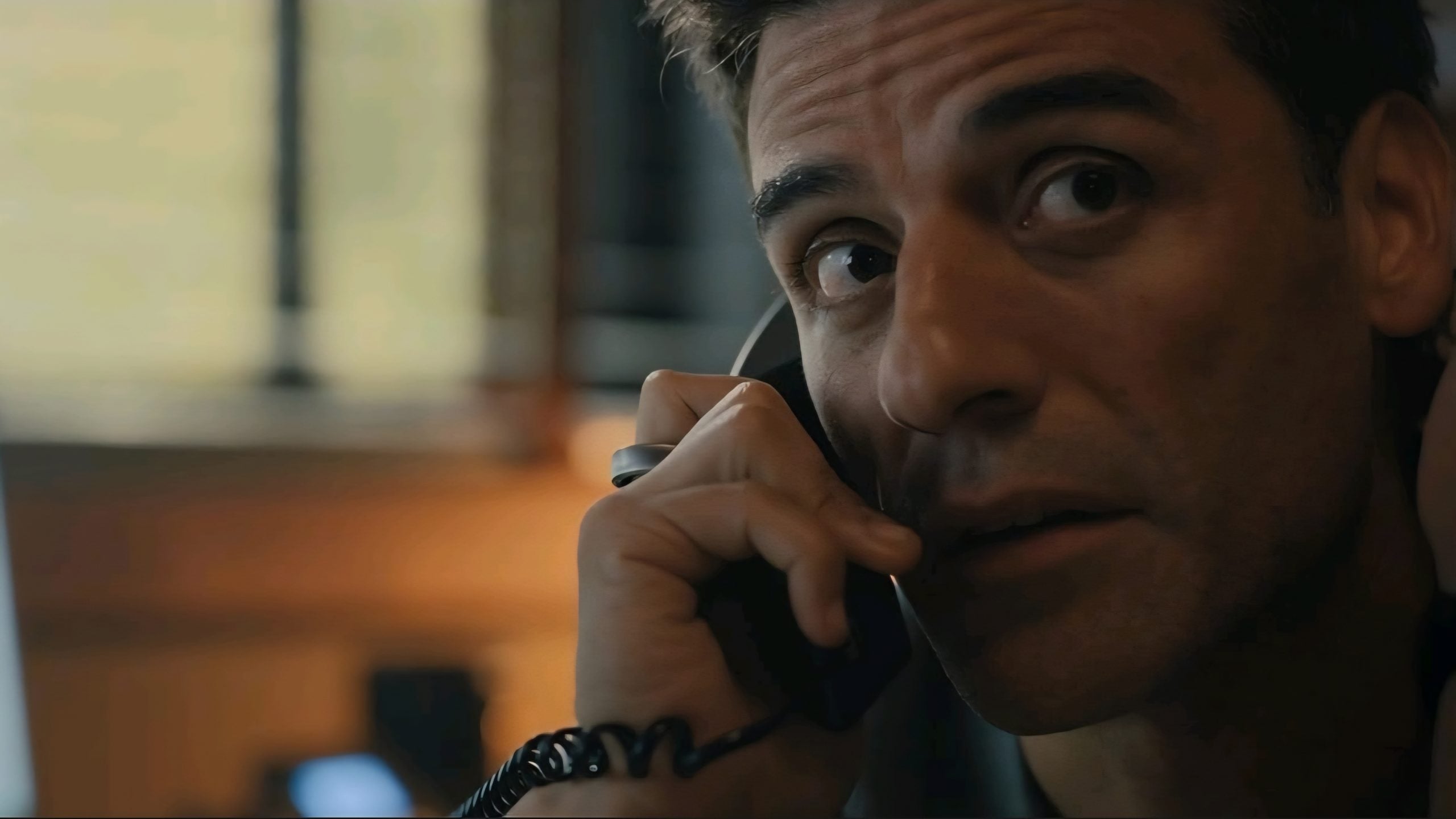 A man holds a phone to his ear, looking upward with a serious expression in a dimly lit room.