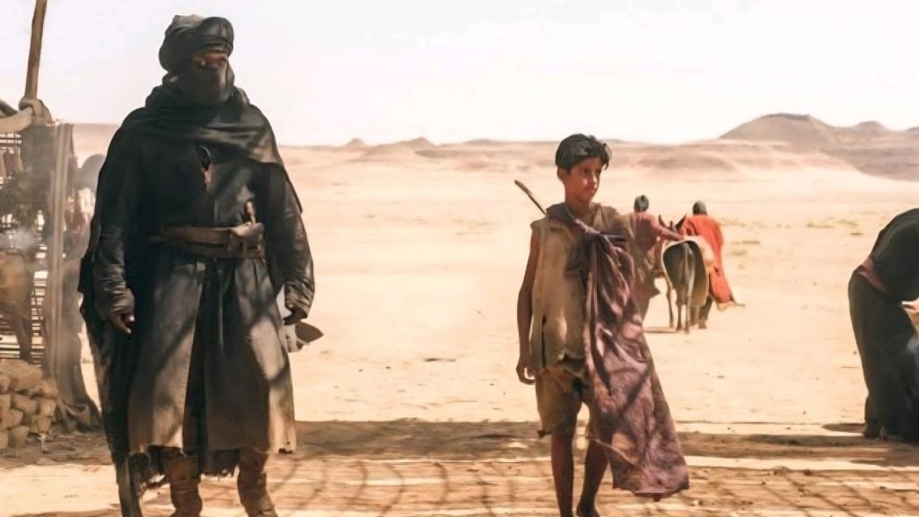 An adult in dark desert clothing walks beside a boy in a tunic, both in a sandy, arid landscape with hills and other figures in the background.