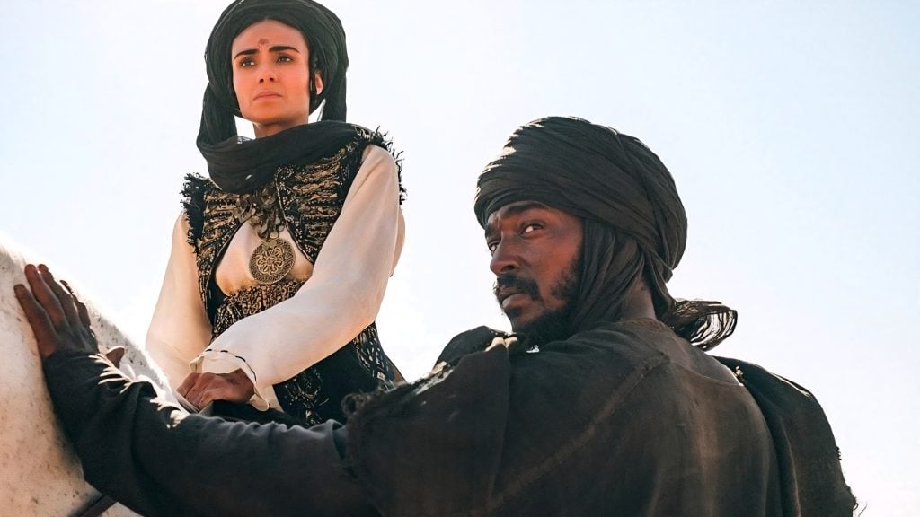 Two people in historical attire and dark headscarves, one seated on horseback and the other standing beside, look into the distance under a clear sky.