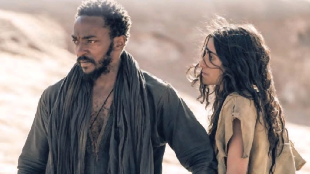 Two people stand outdoors in a barren landscape; the man looks ahead with a serious expression while the woman stands slightly behind him, gazing in the same direction.