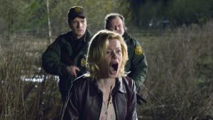 A woman with blonde hair shouting in distress outdoors, accompanied by two law enforcement officers.