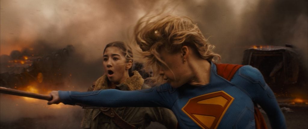 A person in a blue superhero suit with an "S" emblem swings a pole, while another person behind them looks on amid smoke and fire in the background.