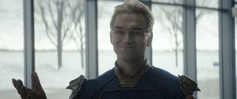 ‘The Boys’ Season 5 Episode 3 Review: Homelander Contends With His Own Mortality A man with blond hair in a blue superhero suit stands indoors, smiling and shrugging, with large windows and snowy trees visible in the background.