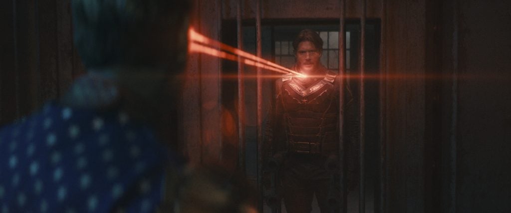 A man in armor stands behind metal bars while another person, partially visible, shoots red laser beams from their eyes toward him.