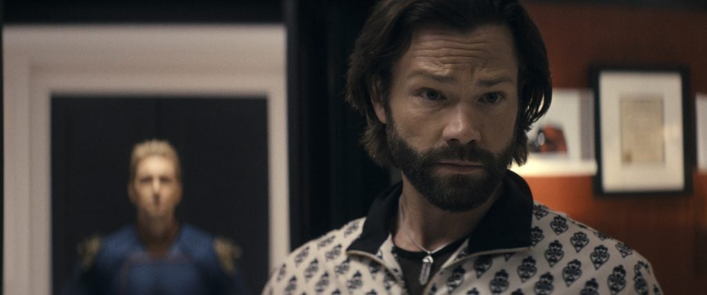 A man with shoulder-length hair and a beard stands in focus indoors, wearing a patterned shirt, while another man in a blue suit appears blurred in the background.