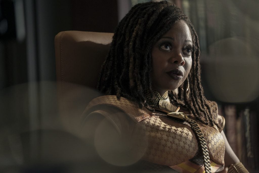 A woman with long, styled hair sits in a chair, wearing a gold outfit and looking slightly upward with a serious expression.