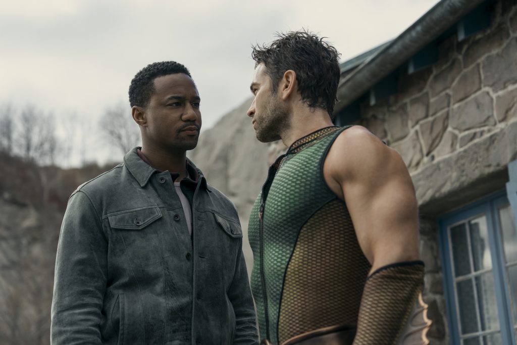 Two men stand face-to-face outside a stone building; one wears a gray jacket, the other is in a sleeveless, textured costume.