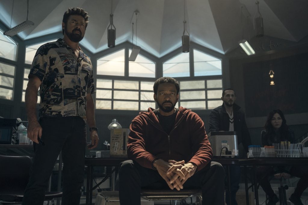 Four people in a laboratory setting, with one man seated in the foreground and three standing behind him, surrounded by scientific equipment and dim lighting.