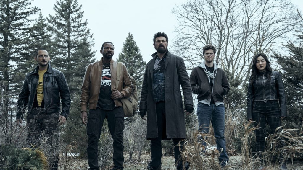 Five people stand outdoors in a wooded area, dressed in casual and rugged clothing, facing forward with serious expressions.