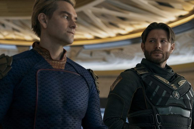 Two men in superhero costumes stand next to each other indoors, looking in different directions with serious expressions.