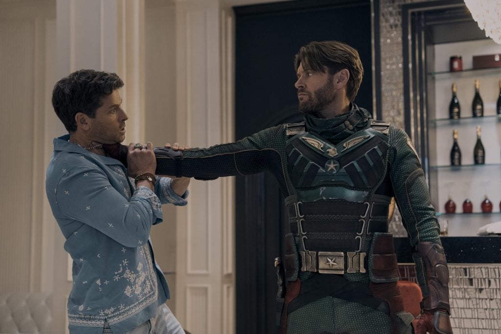 A man in a superhero costume holds another man by the neck with one arm in a modern, upscale room with a display of bottles in the background.