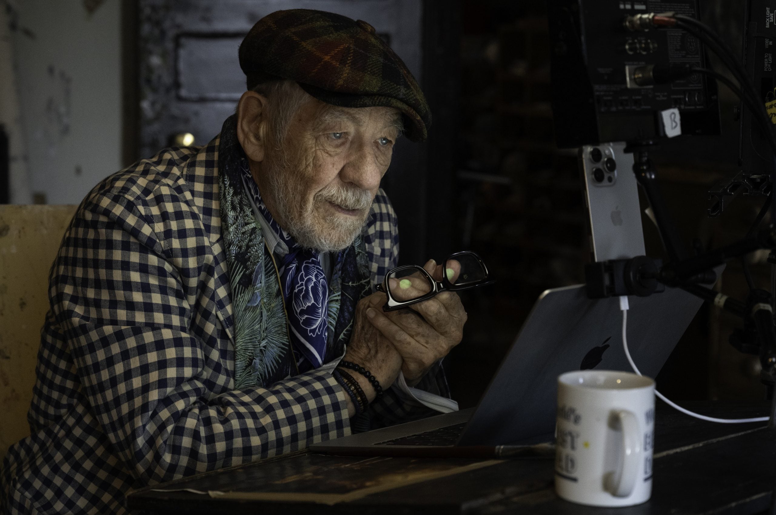 An older man wearing a plaid shirt and cap sits at a table, holding glasses and looking at a laptop screen with a mug and camera equipment nearby.