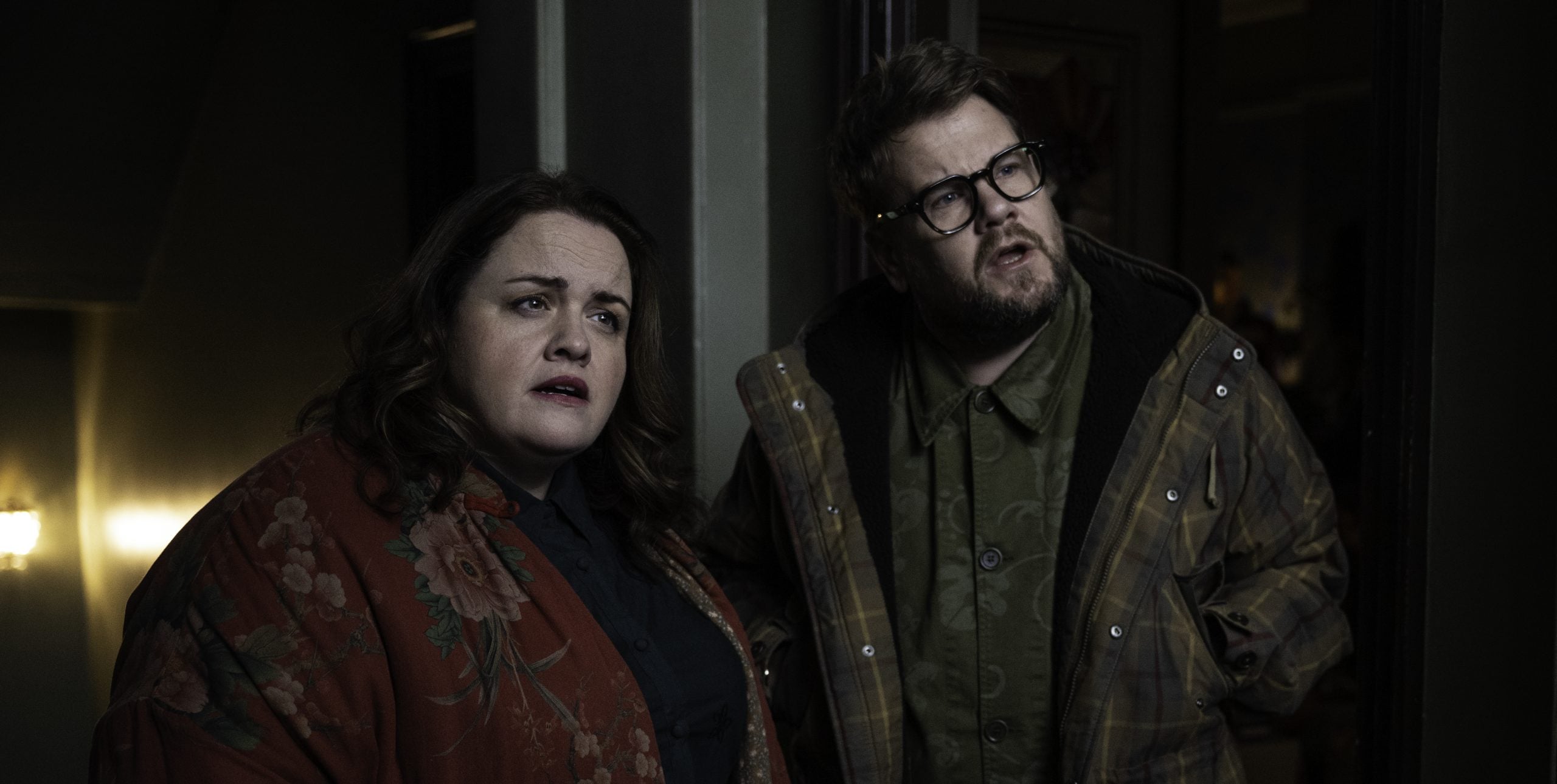 Two people stand indoors, looking in the same direction with concerned expressions. Both wear patterned jackets and glasses. The lighting is dim.