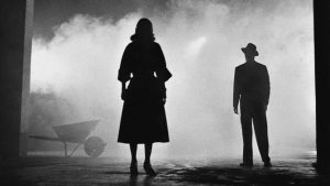 Ignite Films Details New 4K UHD Blu-Ray Release For Film Noir Classic ‘The Big Combo’ Silhouettes of a woman and a man in a foggy, dimly lit setting with a wheelbarrow visible on the left side.