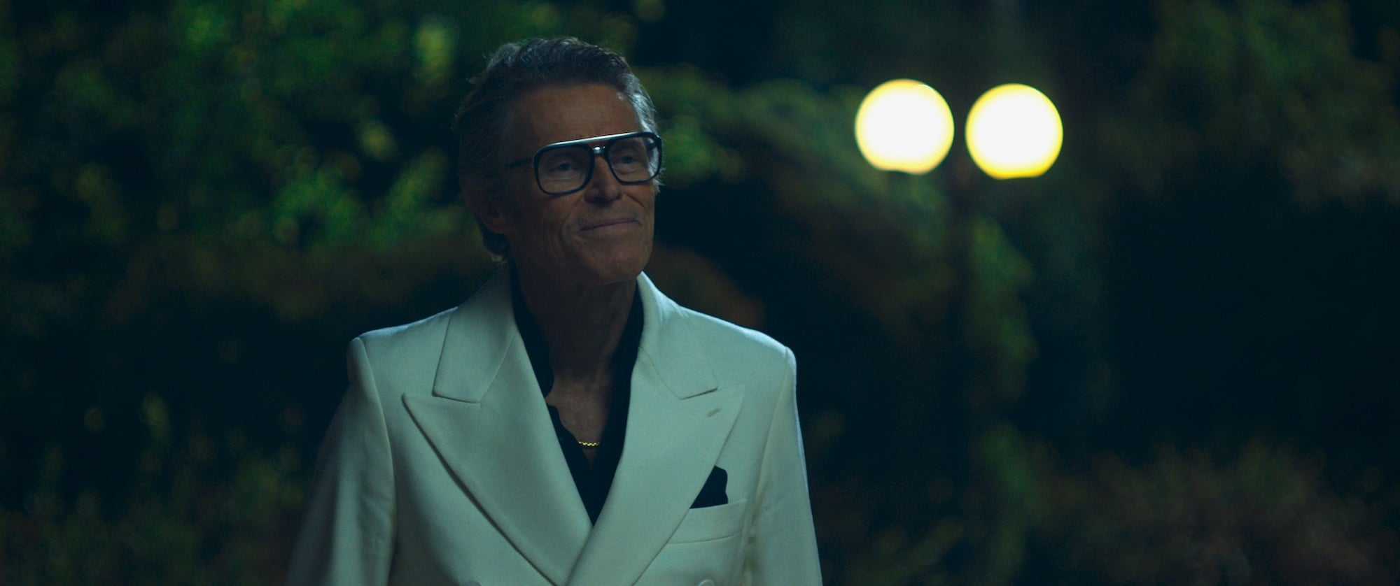 An older man wearing a white double-breasted suit, black shirt, and large glasses stands outdoors at night with blurred lights in the background.