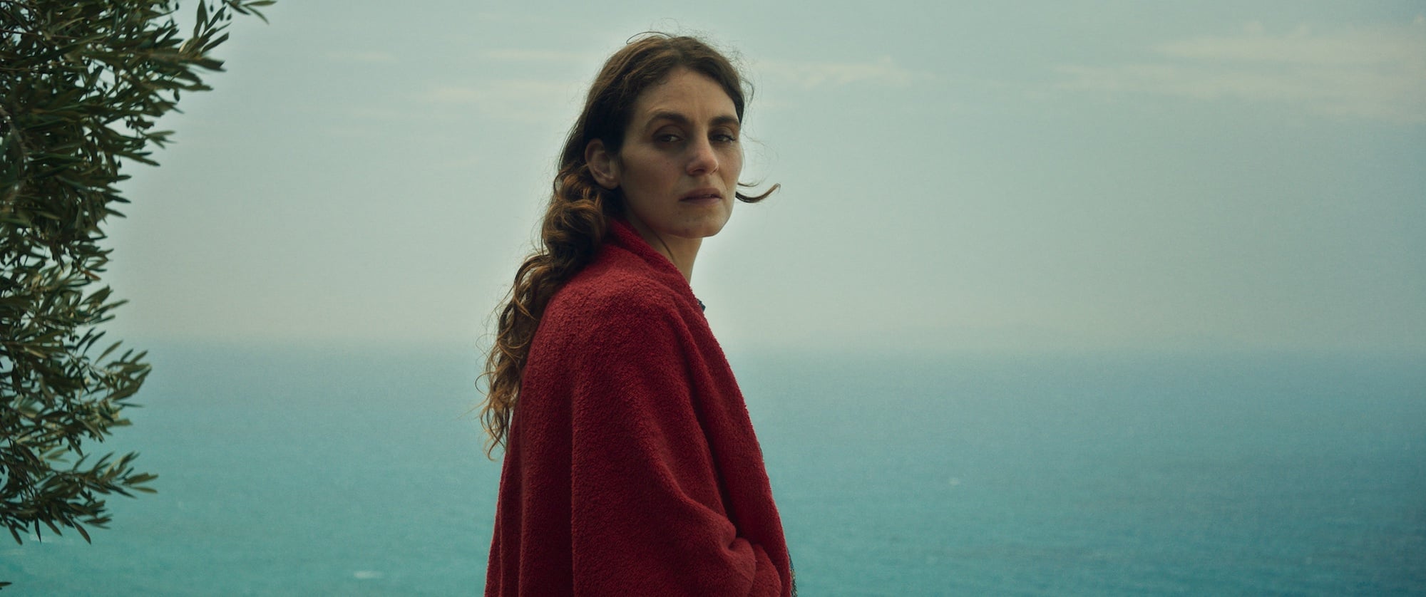A woman in a red coat stands outdoors near a tree, looking at the camera with the ocean and cloudy sky in the background.