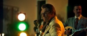 ‘The Birthday Party’ Review – Willem Dafoe Steals The Party In An Otherwise Pedestrian Drama A man in a cream suit stands at a vintage microphone, smiling, with stage lights behind him. Another man in a suit plays a stringed instrument in the background.