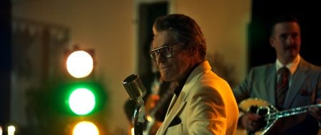 A man in a cream suit stands at a vintage microphone, smiling, with stage lights behind him. Another man in a suit plays a stringed instrument in the background.