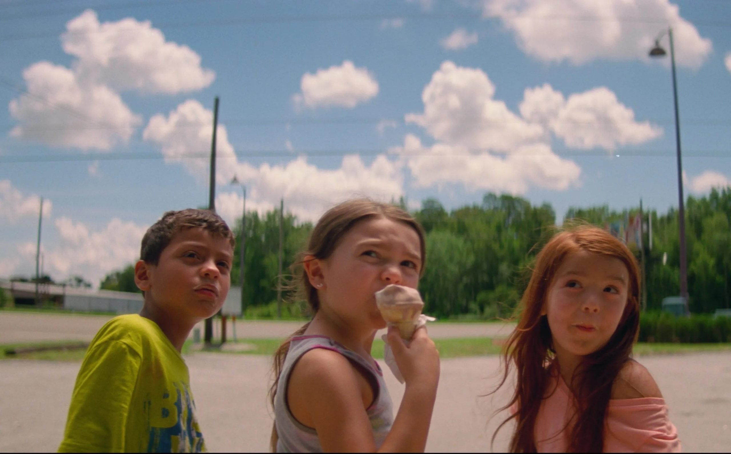 Three children stand outdoors under a partly cloudy sky; the girl in the center eats an ice cream cone while the other two look in different directions.