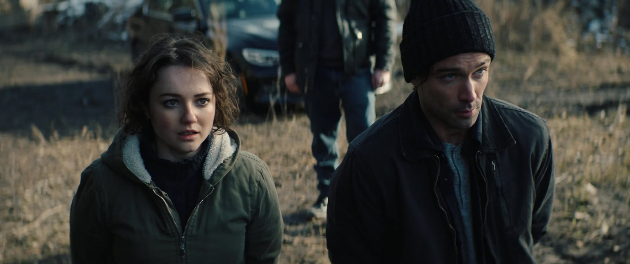 A woman and a man wearing jackets and looking concerned stand outdoors, with another person and a car blurred in the background.