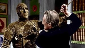 A man in a suit raises a weapon against a mummy-like figure wrapped in aged bandages inside a room with bookshelves and framed art.