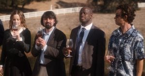 Four people stand outdoors holding glasses of wine, dressed in semi-formal and casual attire, with a white fence and trees in the background.
