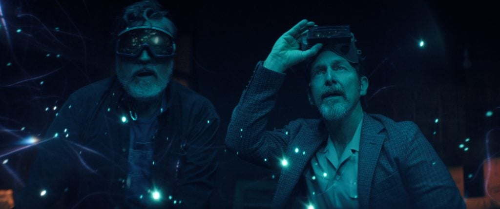Two men wearing augmented reality goggles look up surrounded by floating blue light particles in a dark environment.
