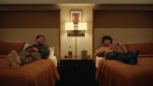 Two people lie on separate twin beds in a dimly lit hotel room, facing each other with a nightstand and lamp between them.