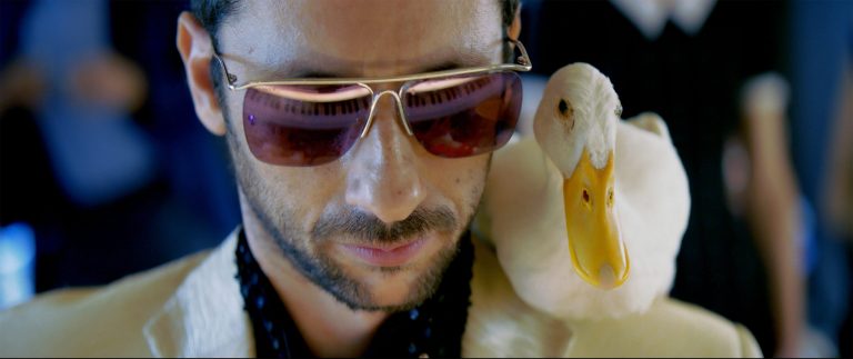 A man wearing sunglasses and a light jacket has a white duck perched on his shoulder.