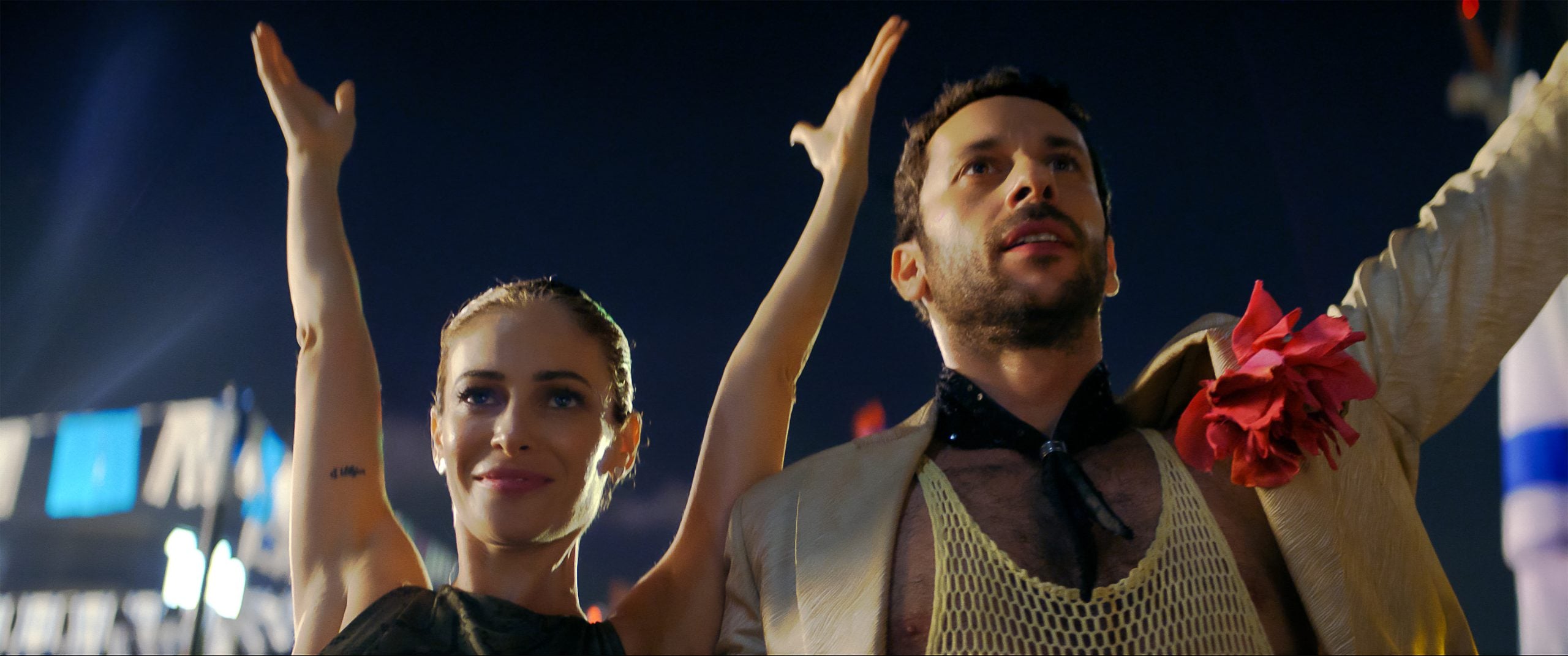 A woman and a man with raised arms stand side by side at night; the man wears a tan jacket with a large pink flower, and both look upward with confident expressions.