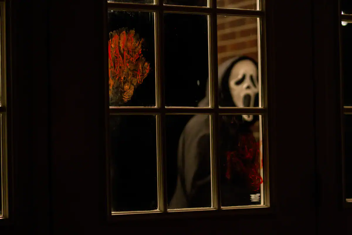 A person in a hood and Ghostface mask stands outside a window at night, partially obscured by glass panes and a red handprint on the glass.