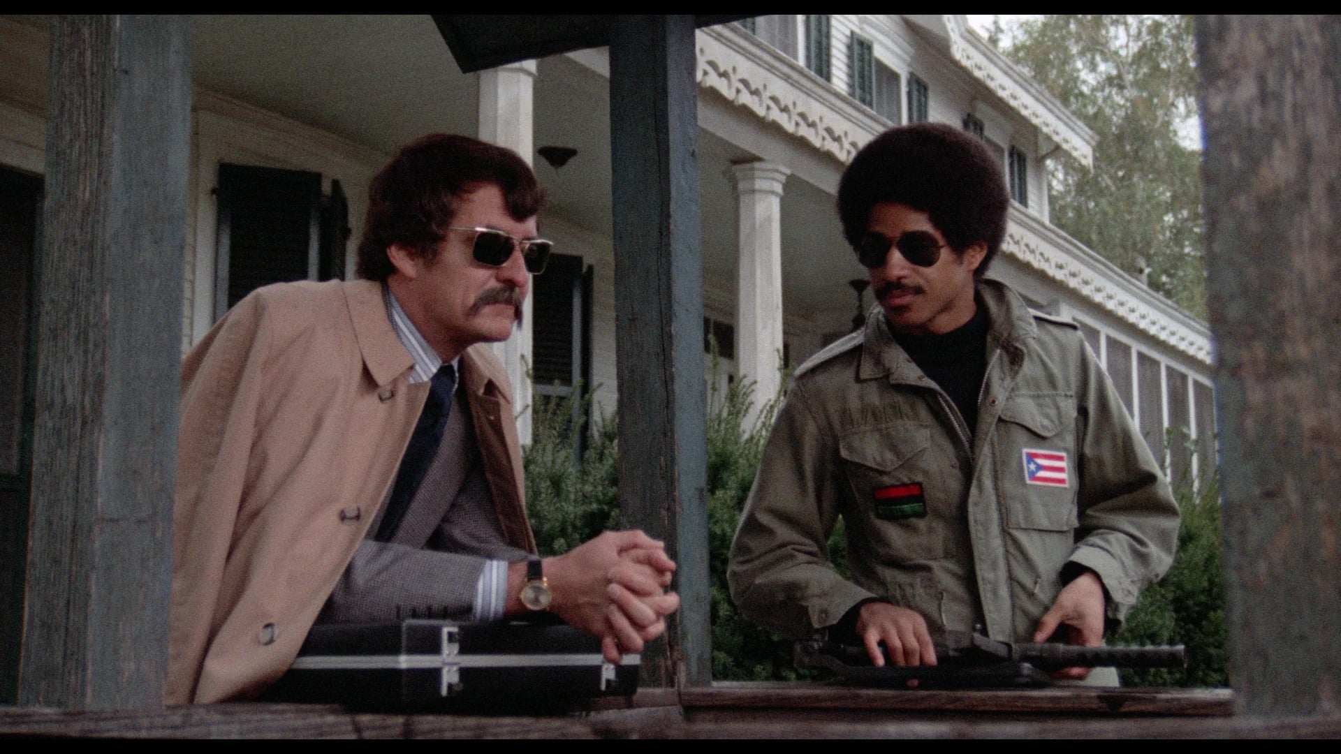 Two men stand at an outdoor wooden structure; one wears a trench coat and sunglasses, the other a military-style jacket with patches and holds a firearm. A large house is visible in the background.