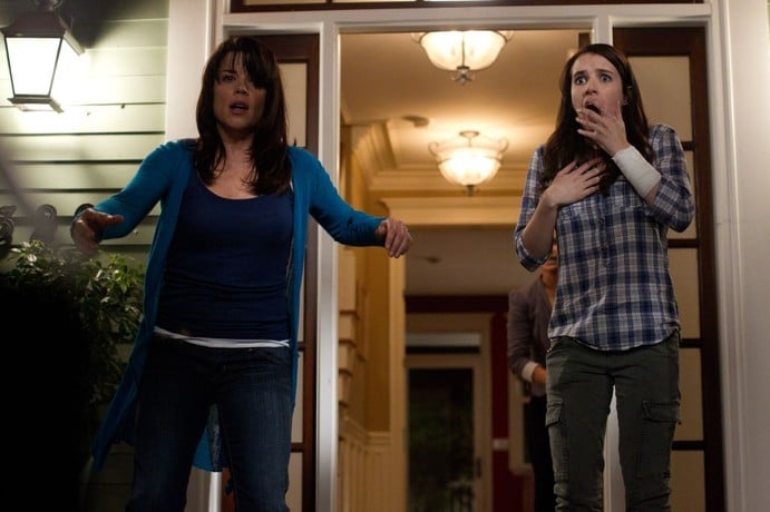 Two women stand outside a house at night; one looks alarmed with arms out, the other covers her mouth in shock. Indoor lights are on behind them.