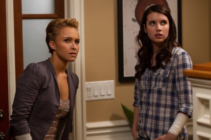 Two young women stand indoors near a light switch, both looking off to the side with serious expressions. One wears a blazer and top, the other a plaid shirt.