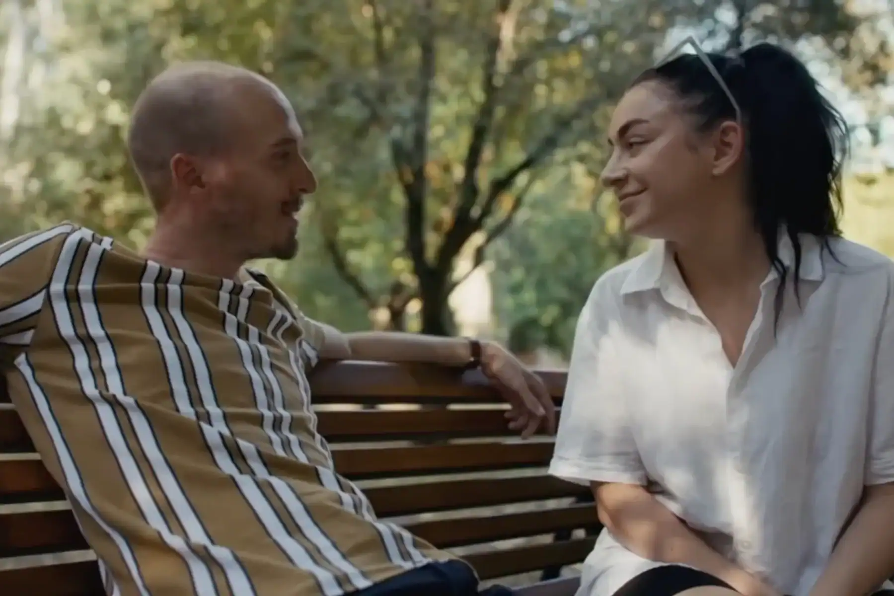 A man and a woman sit on a park bench, facing each other and smiling, with trees in the background.