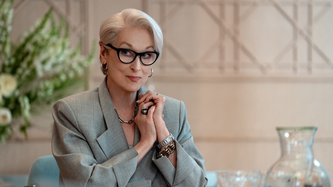 An older woman with short gray hair and glasses, wearing a light gray suit, sits at a table clasping her hands, with flowers and glassware in the background.