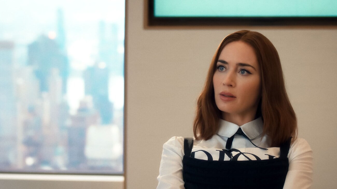 A woman with shoulder-length brown hair sits indoors, wearing a white collared shirt with black detailing, in front of a blurred cityscape window.