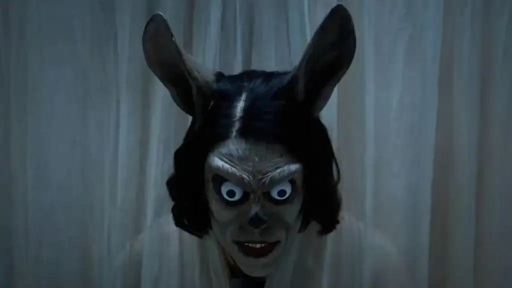 A person with a realistic, creepy rabbit mask featuring large ears and wide, intense eyes stands in front of sheer white curtains.