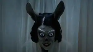 A person with a realistic, creepy rabbit mask featuring large ears and wide, intense eyes stands in front of sheer white curtains.