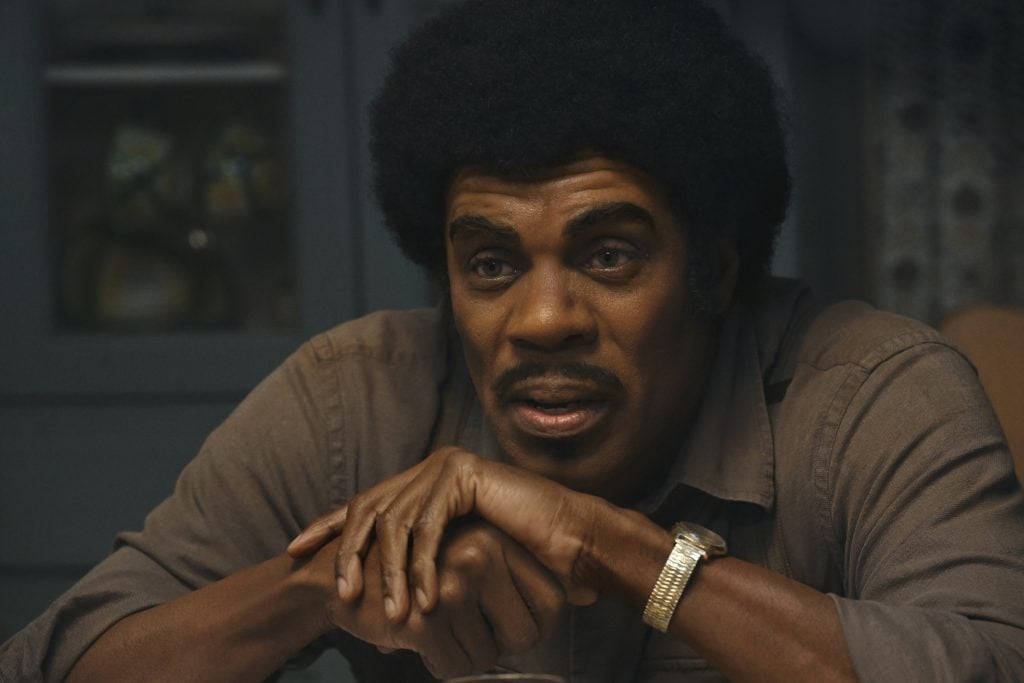 A man with an afro hairstyle and mustache sits at a table, resting his arms on the surface, wearing a brown shirt and a gold wristwatch.