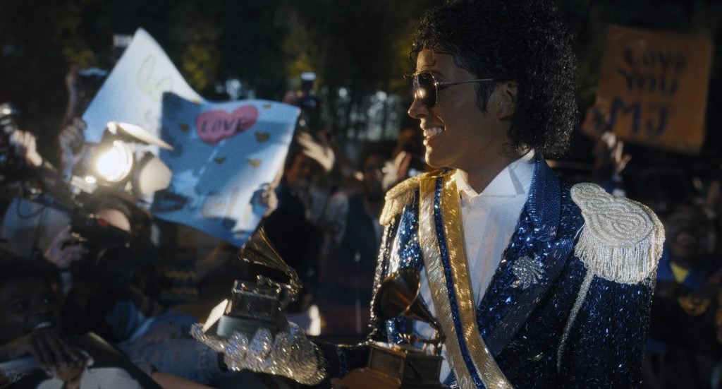 A person in a blue sequined jacket and sunglasses holds Grammy awards while smiling at a crowd of fans holding signs and cameras.