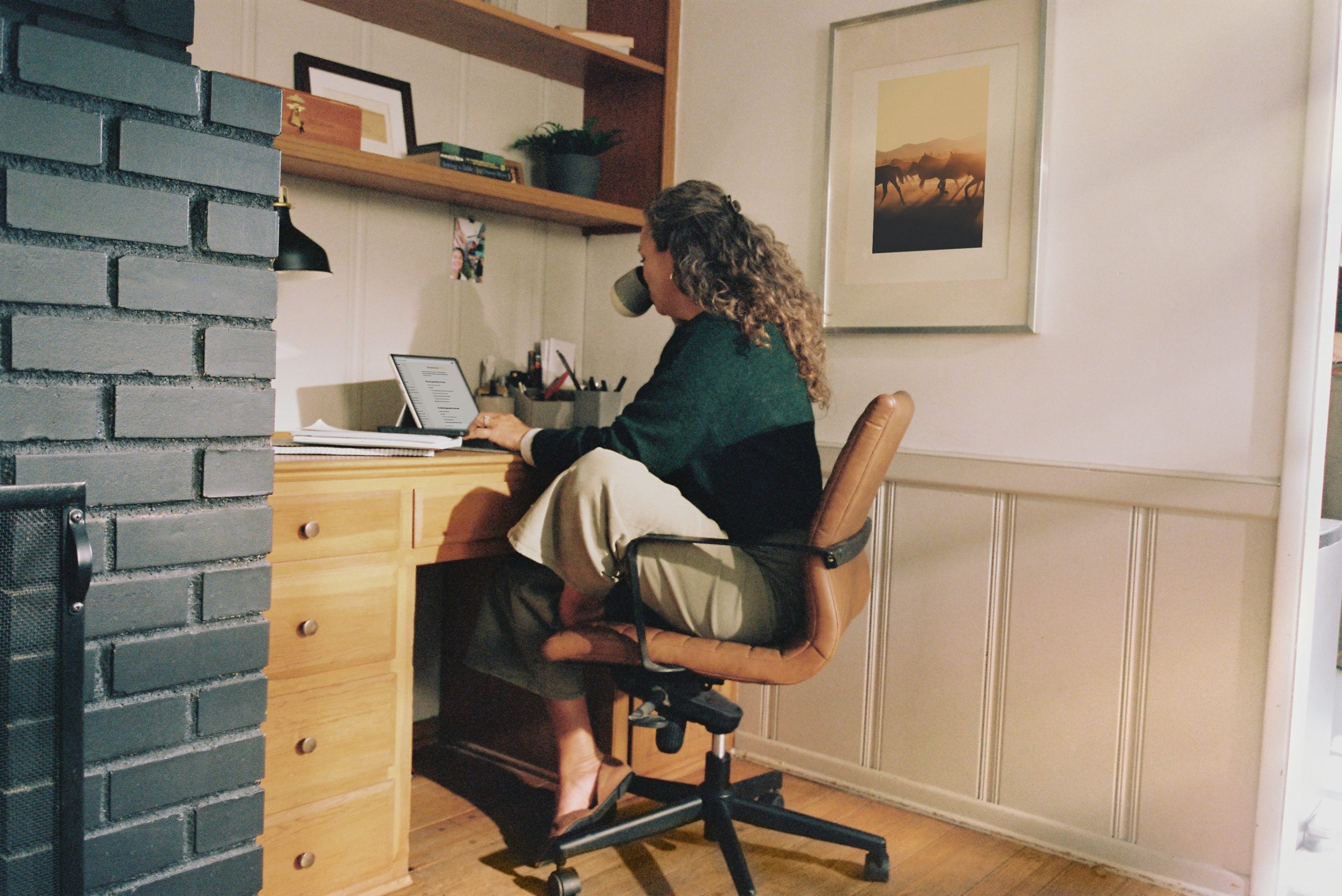 A person sits at a wooden desk, working on a laptop and drinking from a mug in a home office with shelves and framed artwork on the wall.