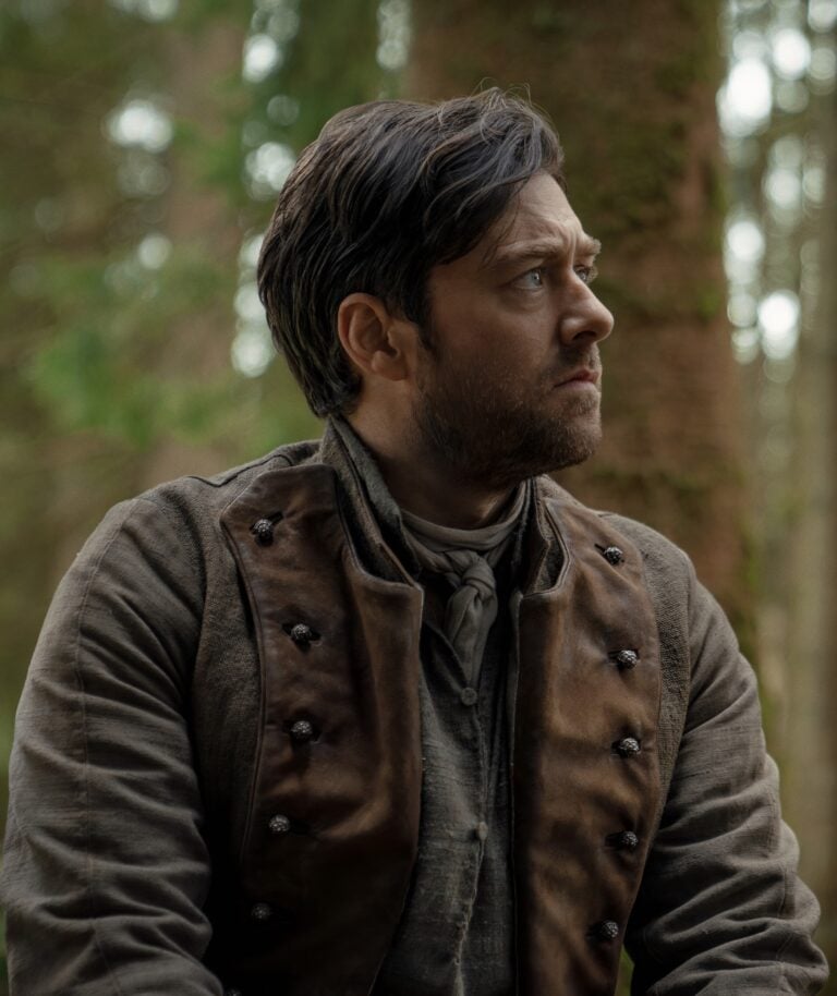 A man in historical clothing with dark hair and a beard looks to the side while sitting outdoors in a forest.